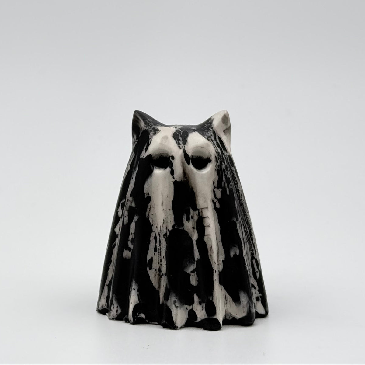 A handmade ghost cat figurine by The Nordic Ghost Merchants. The ghost ornament is bone white with a black, bark like pattern. Front view on a grey studio background.