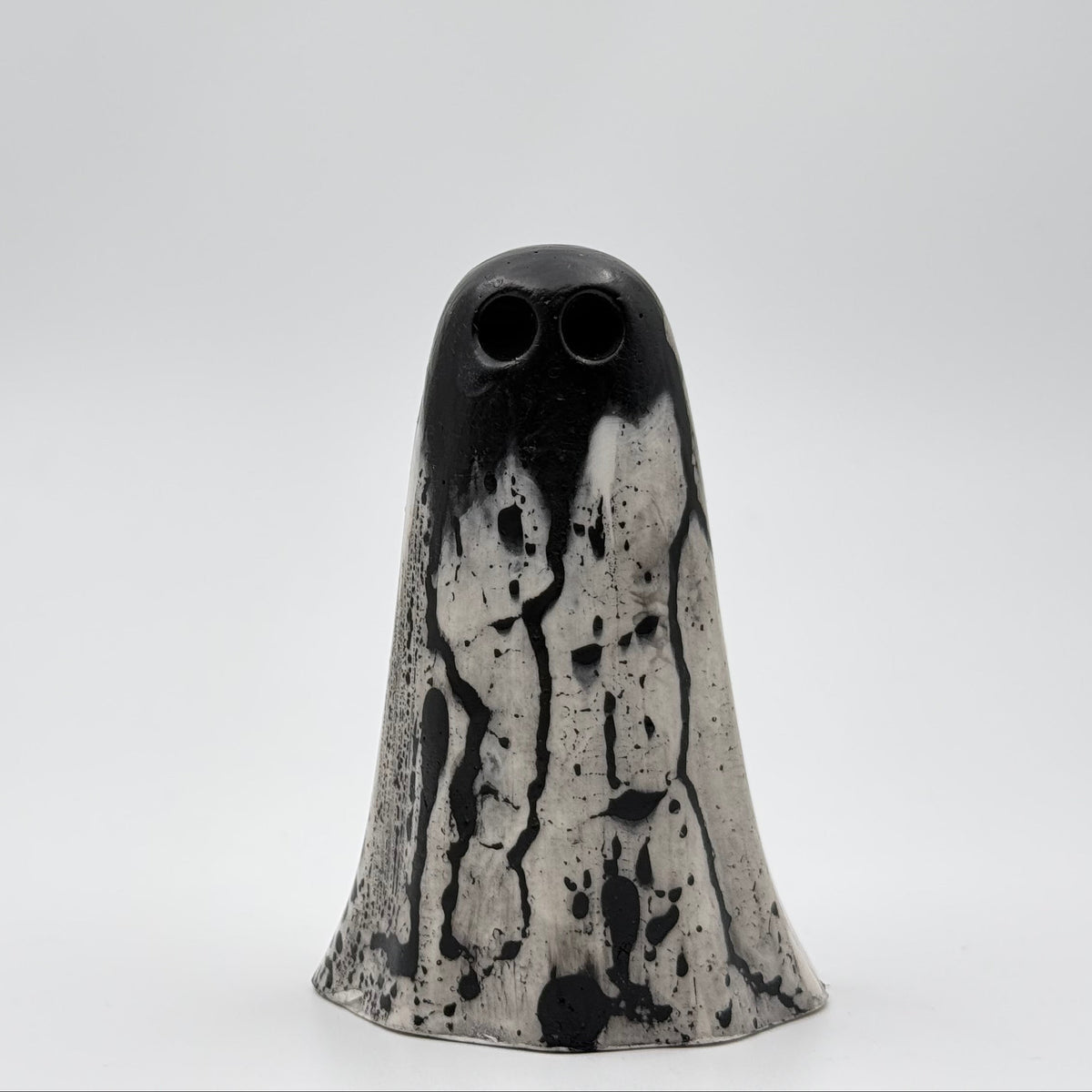 A handmade ghost figurine made by The Nordic Ghost Merchants in Denmark. The ghost is bone white with black, bark like patterning. Front view on a grey studio background.