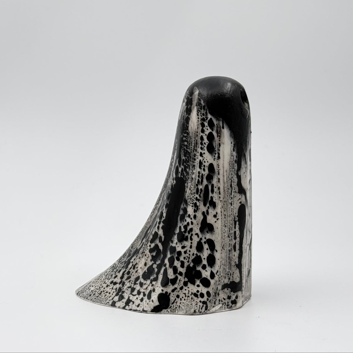 A handmade ghost figurine made by The Nordic Ghost Merchants in Denmark. The ghost is bone white with black, bark like patterning. Side view on a grey studio background.
