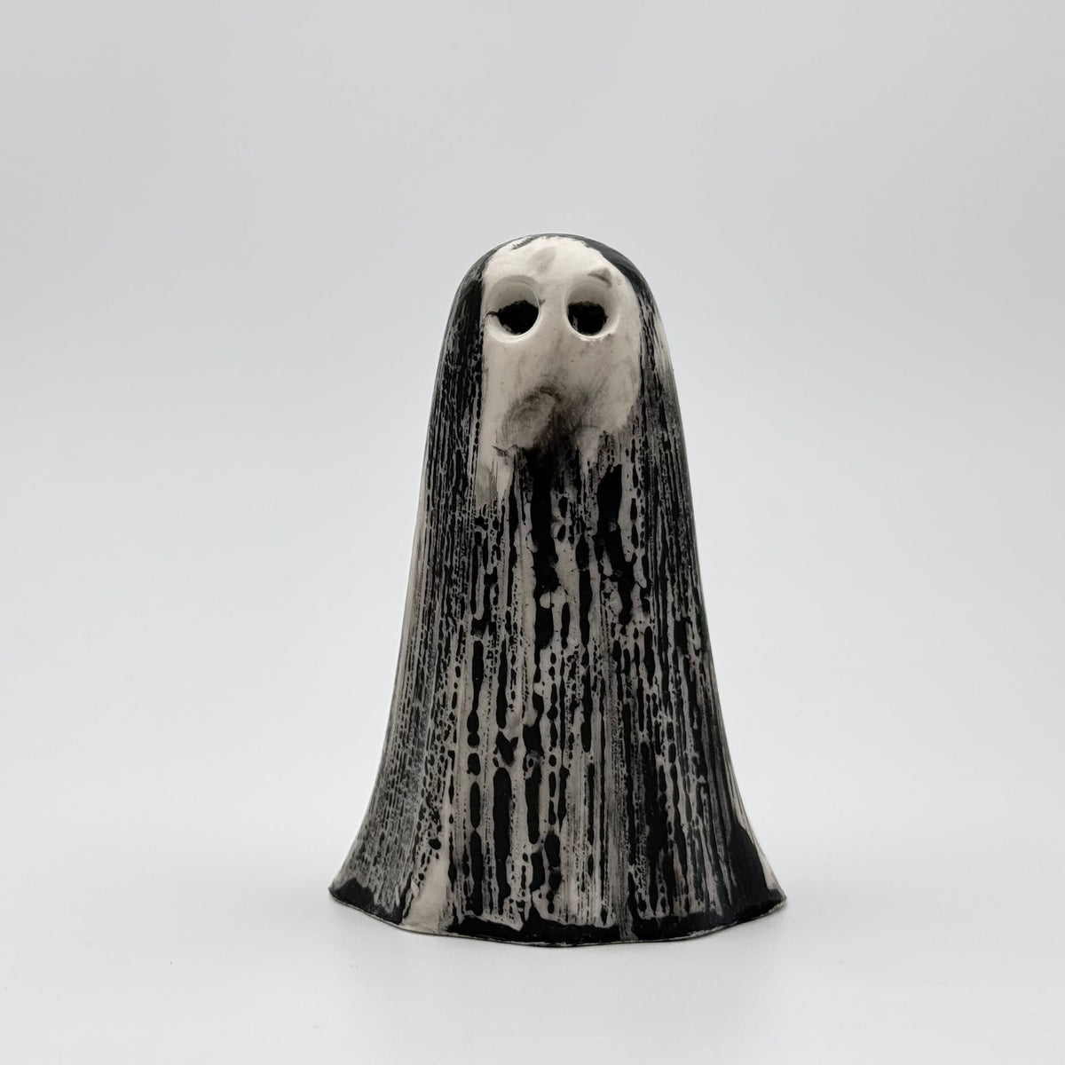 A handmade ghost figurine made by The Nordic Ghost Merchants. The ghost is bone white with black, bark like patterning. Front view on a grey studio background.