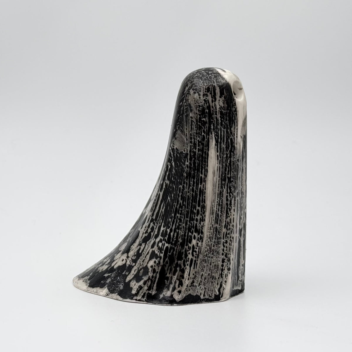A handmade ghost figurine made by The Nordic Ghost Merchants. The ghost is bone white with black, bark like patterning. Side view on a grey studio background.