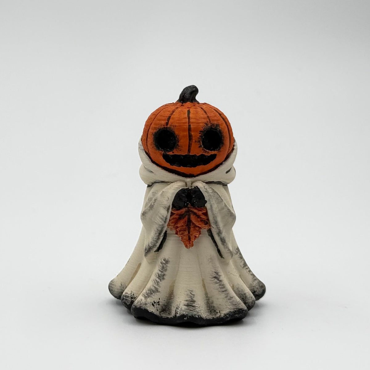 A hand painted folklore figurine made by The Nordic Ghost Merchants in Denmark. The figurine has a carved pumpkin head and is robed in cream cloth while he clutches an autumn leaf. Front view on a grey studio background.