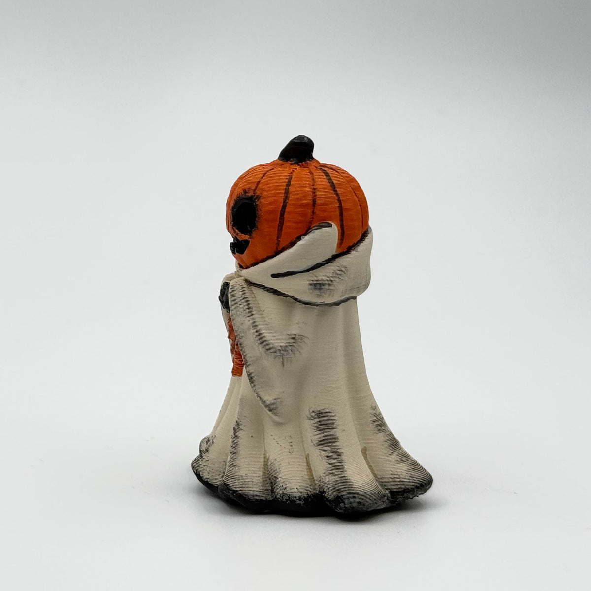 A hand painted folklore figurine made by The Nordic Ghost Merchants in Denmark. The figurine has a carved pumpkin head and is robed in cream cloth while he clutches an autumn leaf. Side view on a grey studio background.