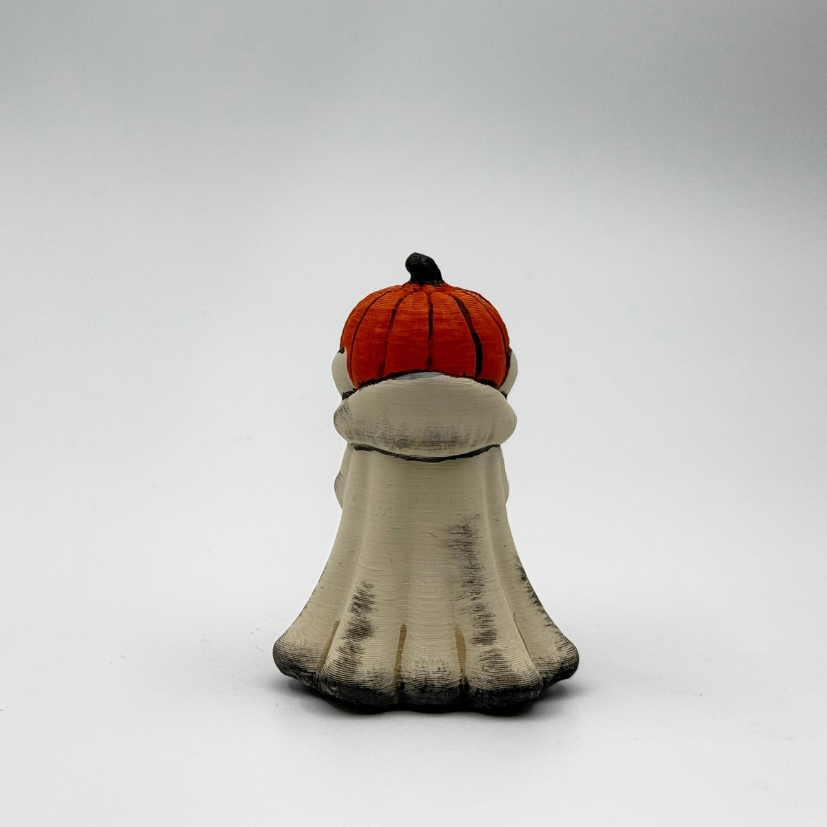 A hand painted folklore figurine made by The Nordic Ghost Merchants in Denmark. The figurine has a carved pumpkin head and is robed in cream cloth while he clutches an autumn leaf. Rear view on a grey studio background.