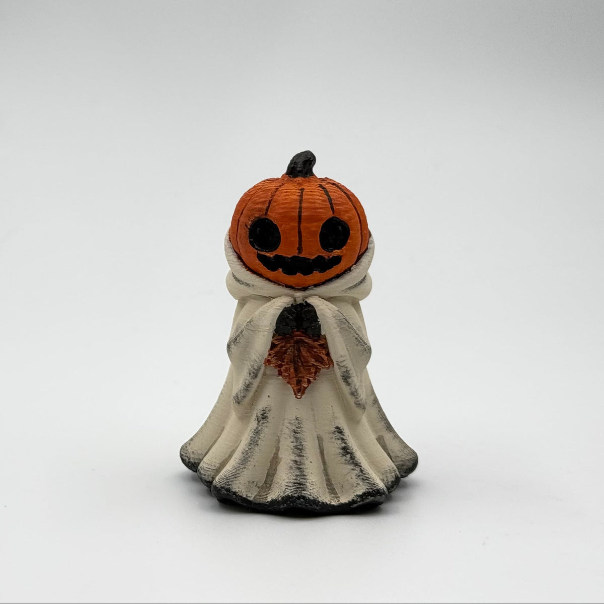 A hand painted folklore figurine made by The Nordic Ghost Merchants in Denmark. The figurine has a carved pumpkin head and is robed in cream cloth while he clutches an autumn leaf. Front view on a grey studio background.