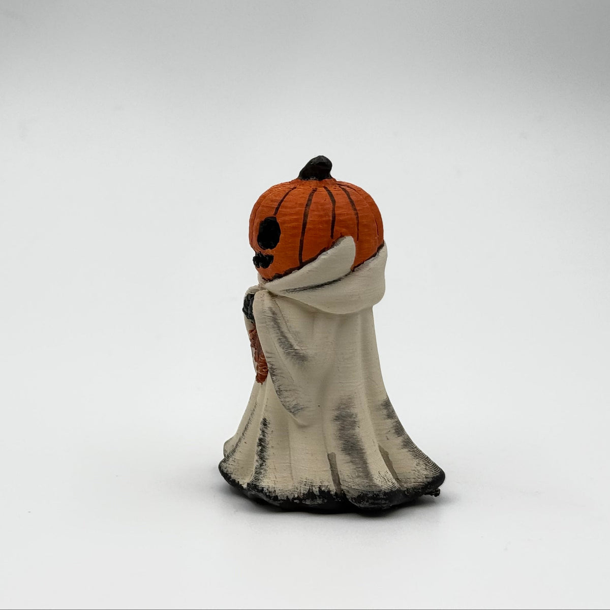 A hand painted folklore figurine made by The Nordic Ghost Merchants in Denmark. The figurine has a carved pumpkin head and is robed in cream cloth while he clutches an autumn leaf. Side view on a grey studio background.