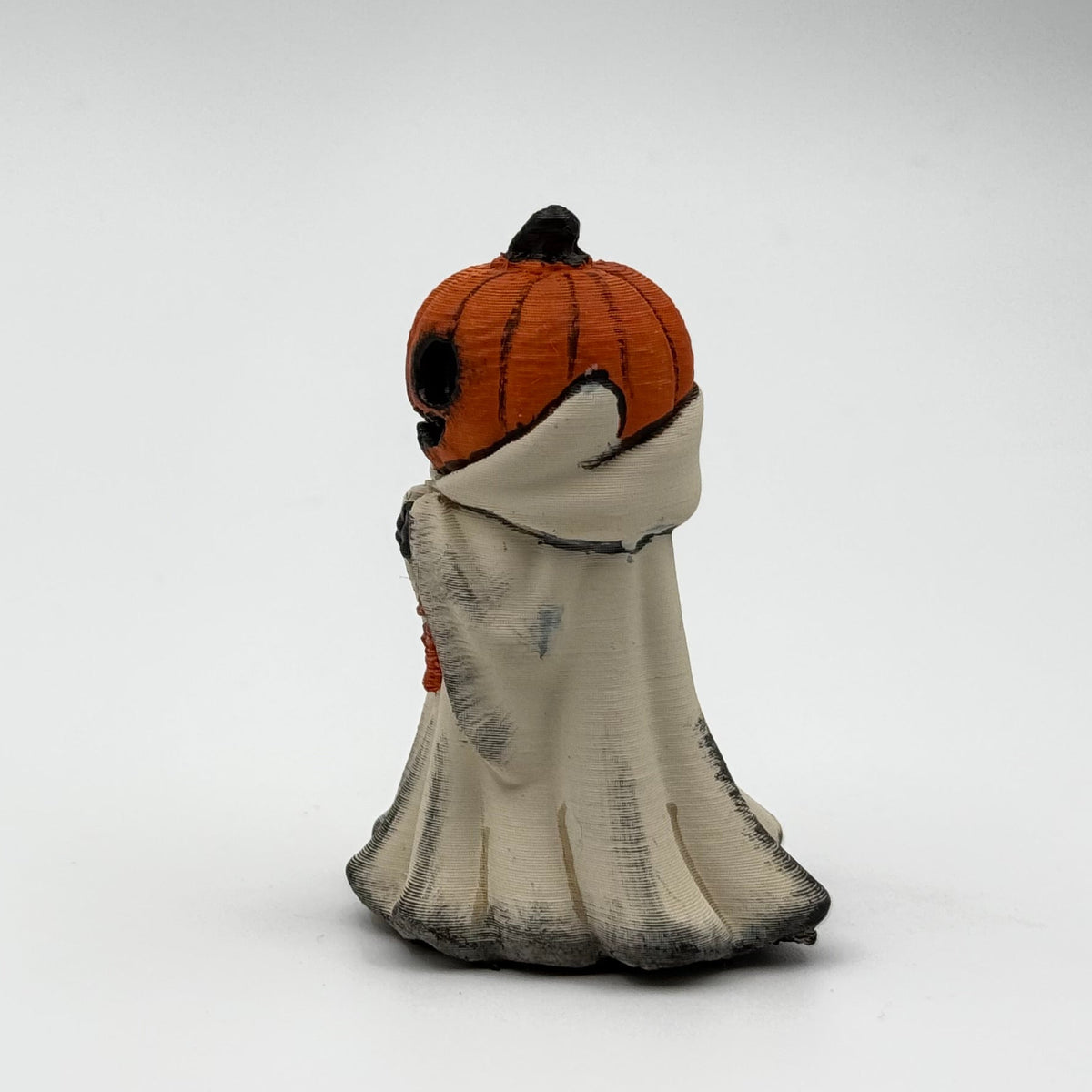 A hand painted folklore figurine made by The Nordic Ghost Merchants in Denmark. The figurine has a carved pumpkin head and is robed in cream cloth while he clutches an autumn leaf. Side view on a grey studio background.