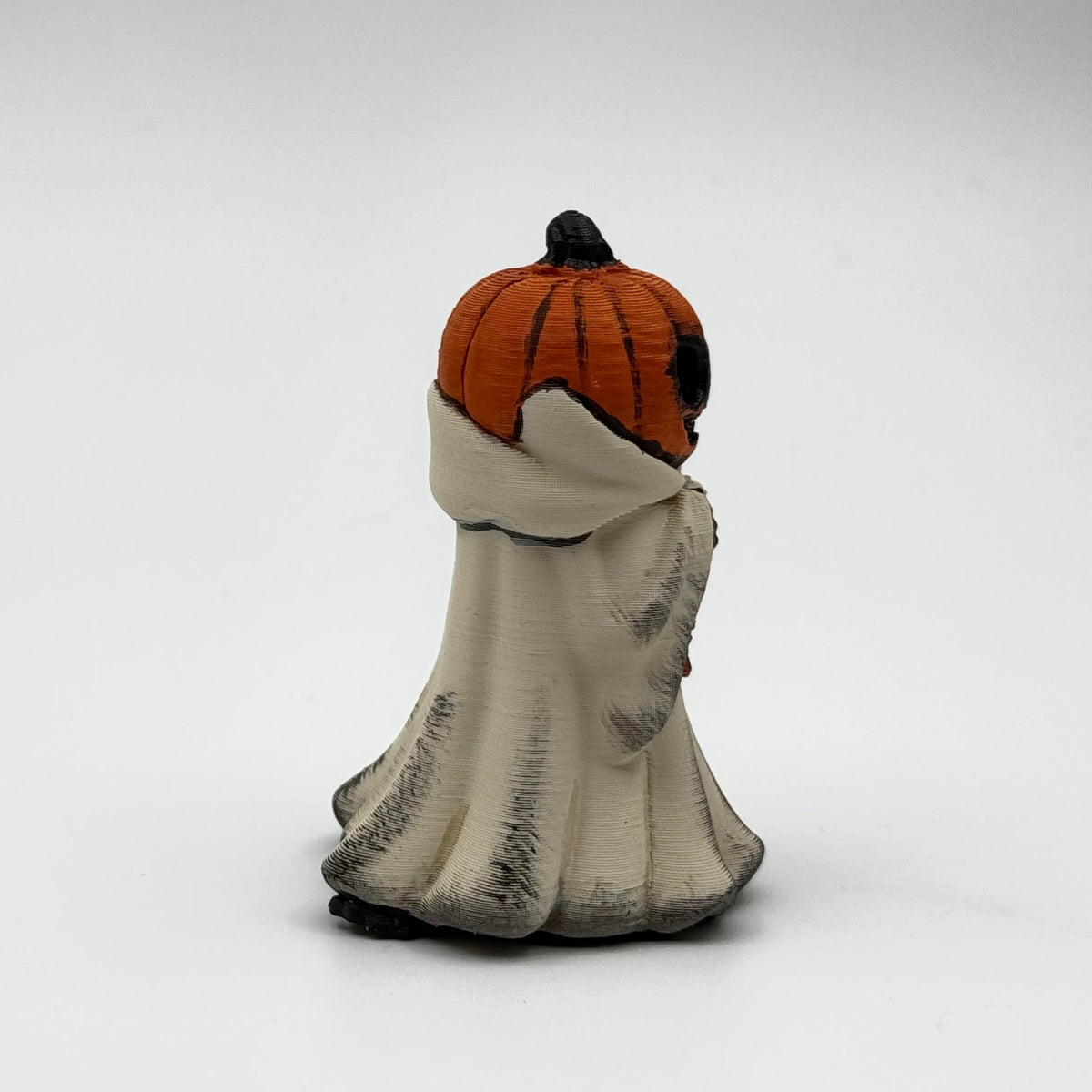A hand painted folklore figurine made by The Nordic Ghost Merchants in Denmark. The figurine has a carved pumpkin head and is robed in cream cloth while he clutches an autumn leaf. Side view on a grey studio background.