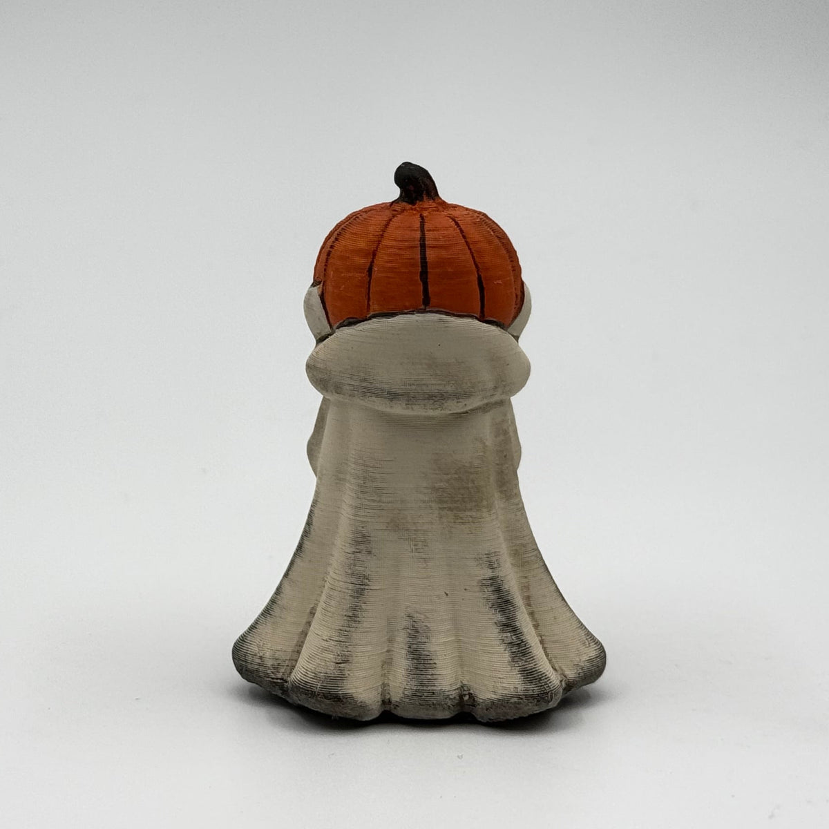 A hand painted folklore figurine made by The Nordic Ghost Merchants in Denmark. The figurine has a carved pumpkin head and is robed in cream cloth while he clutches an autumn leaf. Rear view on a grey studio background.