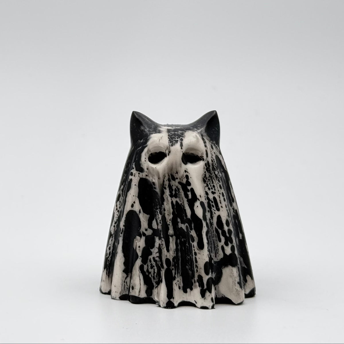 A handmade ghost cat figurine made by The Nordic Ghost Merchants. The ghost ornament is bone white with black, bark like patterning. Front view on a grey studio background.