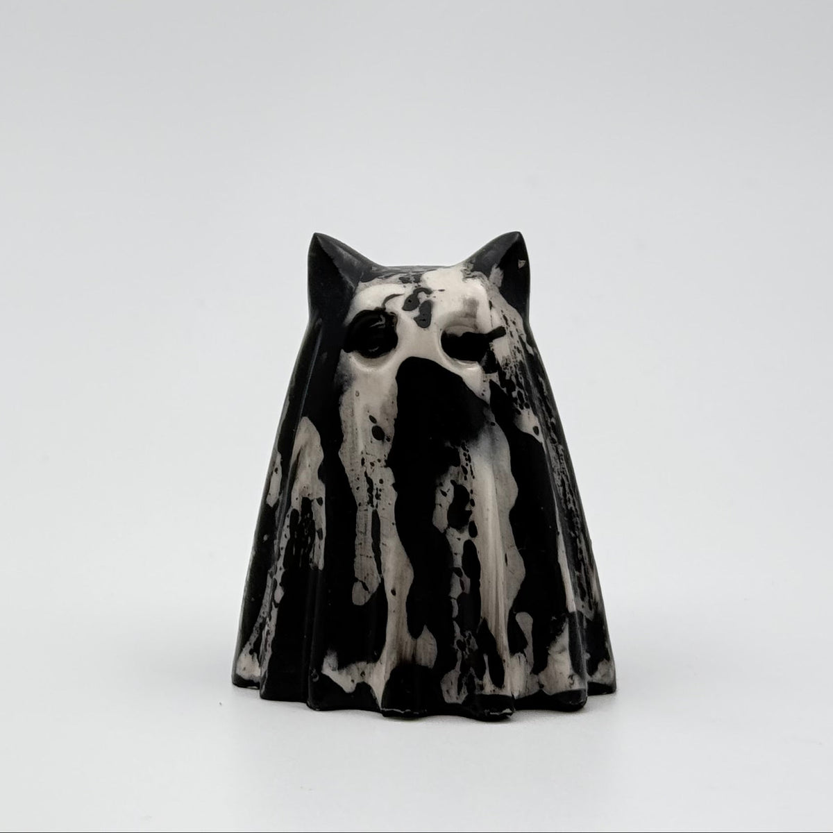 A handmade ghost cat figurine by The Nordic Ghost Merchants. The ghost ornament is bone white with black, bark like patterning. Front view on a grey studio background.