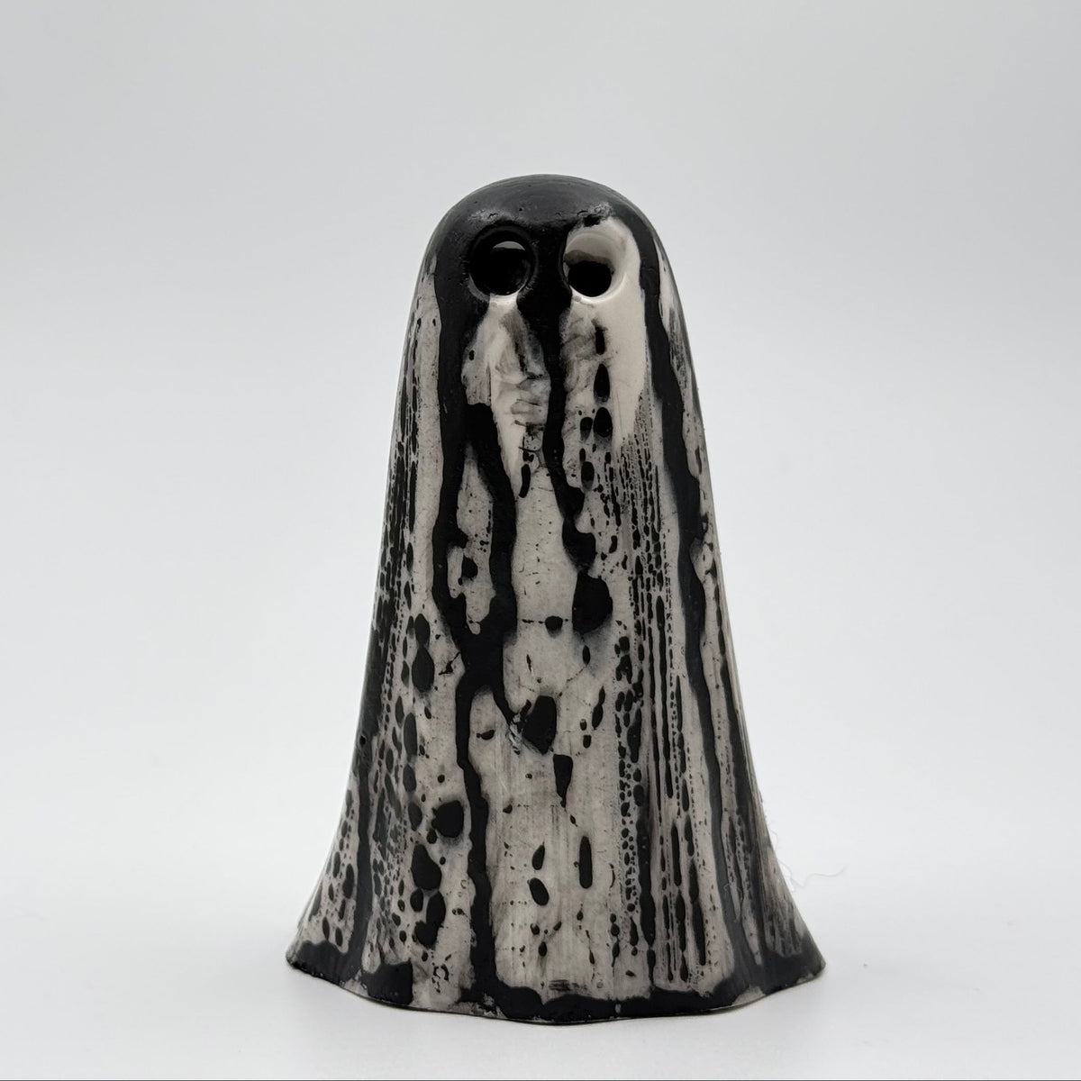 A handmade ghost figurine made by The Nordic Ghost Merchants. The base of the ghost is bone white and it has black, bark like patterning over the full body of the ghost ornament. Front view against a grey studio background.