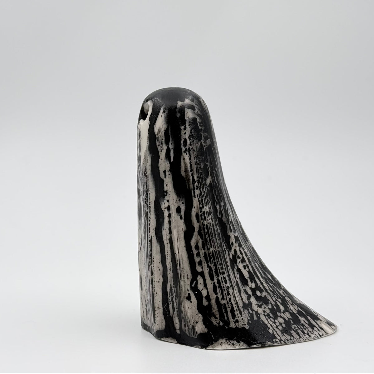A handmade ghost figurine made by The Nordic Ghost Merchants. The base of the ghost is bone white and it has black, bark like patterning over the full body of the ghost ornament. Side view against a grey studio background.