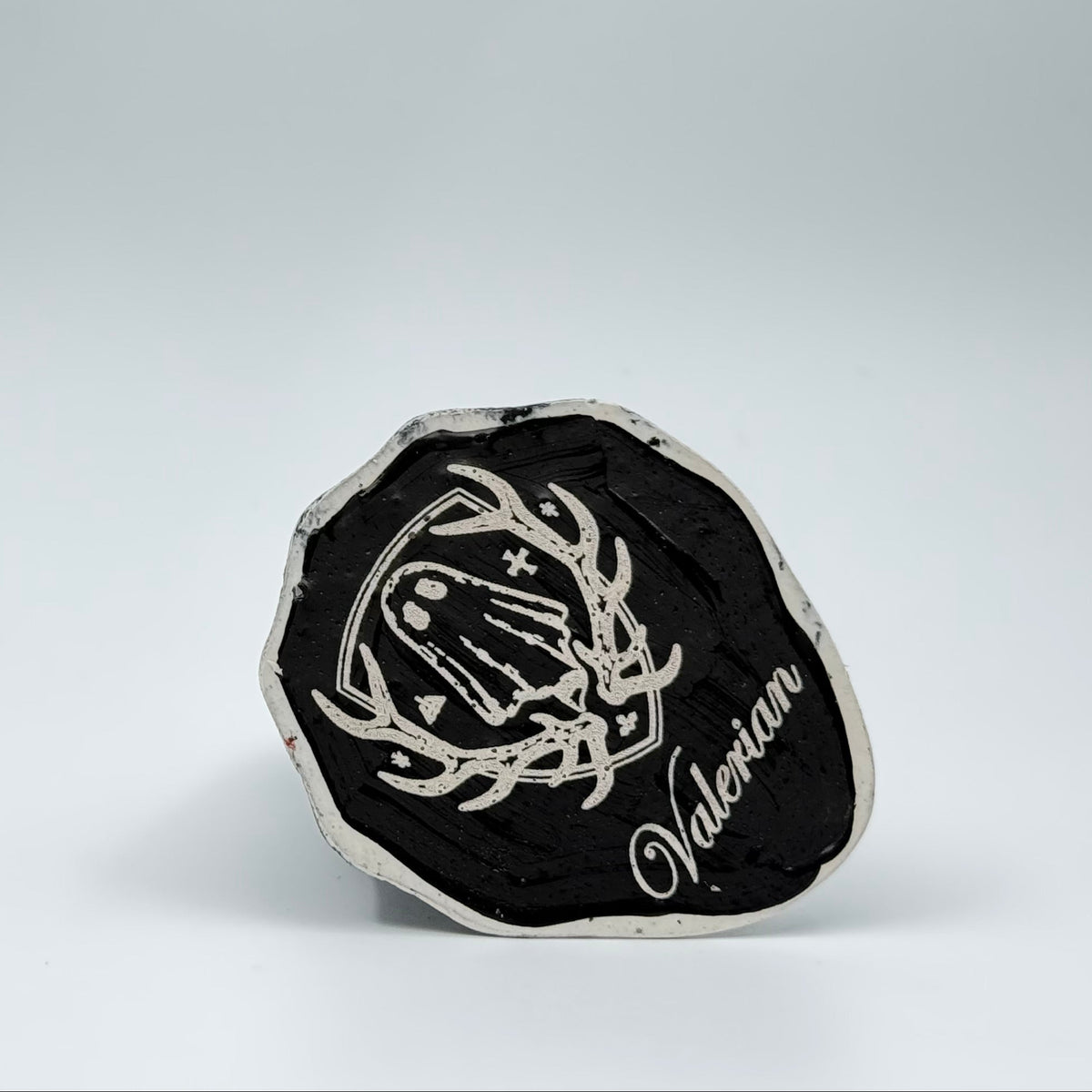 A handmade ghost figurine by The Nordic Ghost Merchants. The view is of the base of the ghost ornament, painted black with the brand logo engraved on it - a coat of arms with a sheet ghost in the centre.
