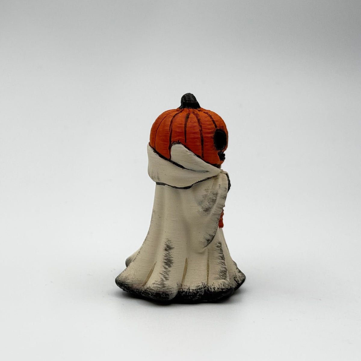 A hand painted folklore figurine made by The Nordic Ghost Merchants in Denmark. The figurine has a carved pumpkin head and is robed in cream cloth while he clutches an autumn leaf. Side view on a grey studio background.