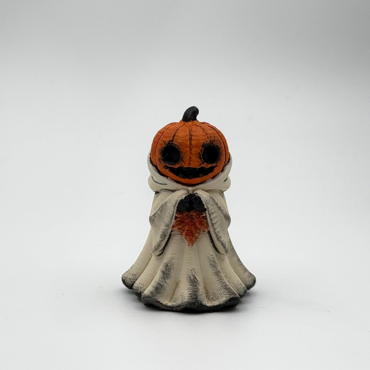 A hand painted folklore figurine made by The Nordic Ghost Merchants in Denmark. The figurine has a carved pumpkin head and is robed in cream cloth while he clutches an autumn leaf. Front view on a grey studio background.
