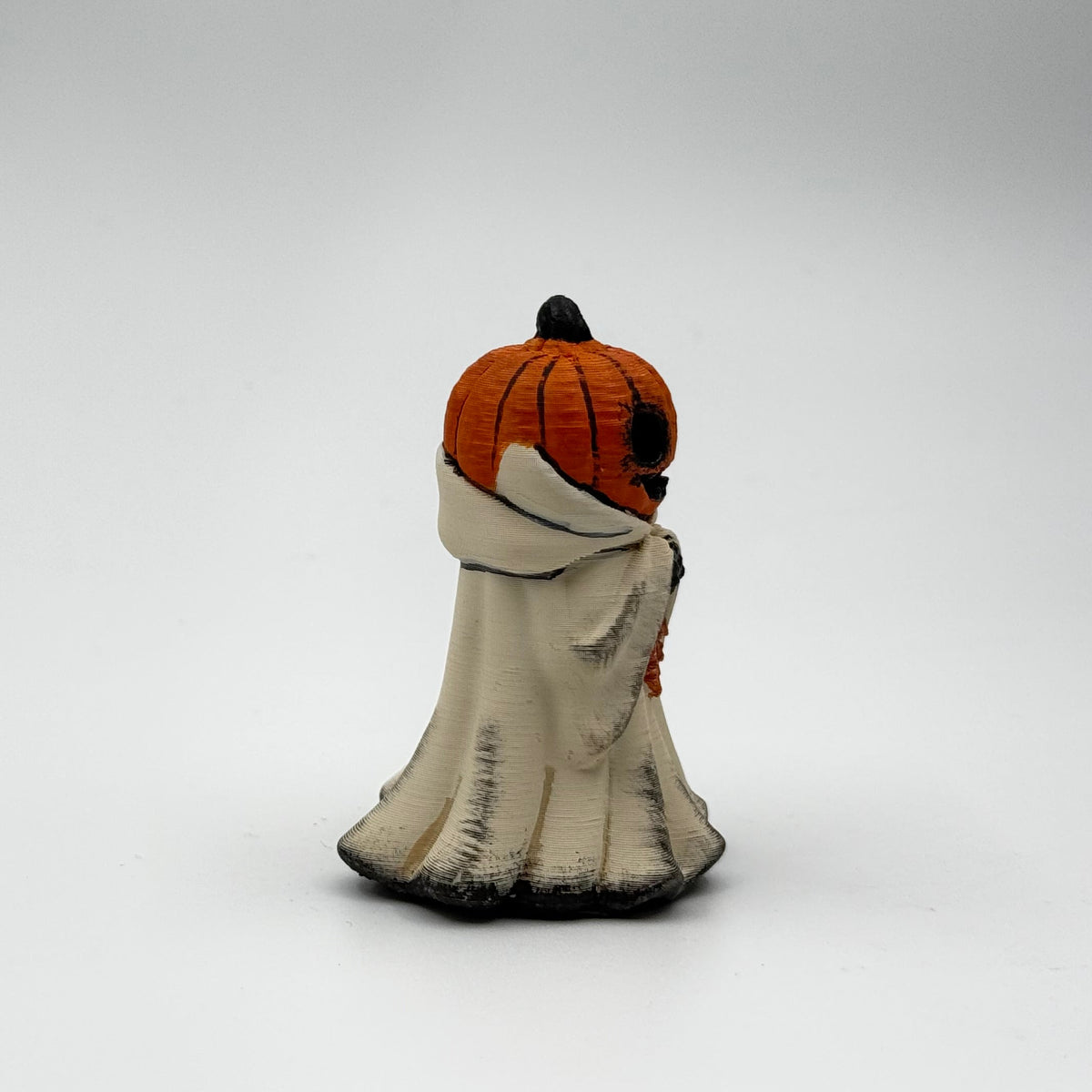 A hand painted folklore figurine made by The Nordic Ghost Merchants in Denmark. The figurine has a carved pumpkin head and is robed in cream cloth while he clutches an autumn leaf. Side view on a grey studio background.