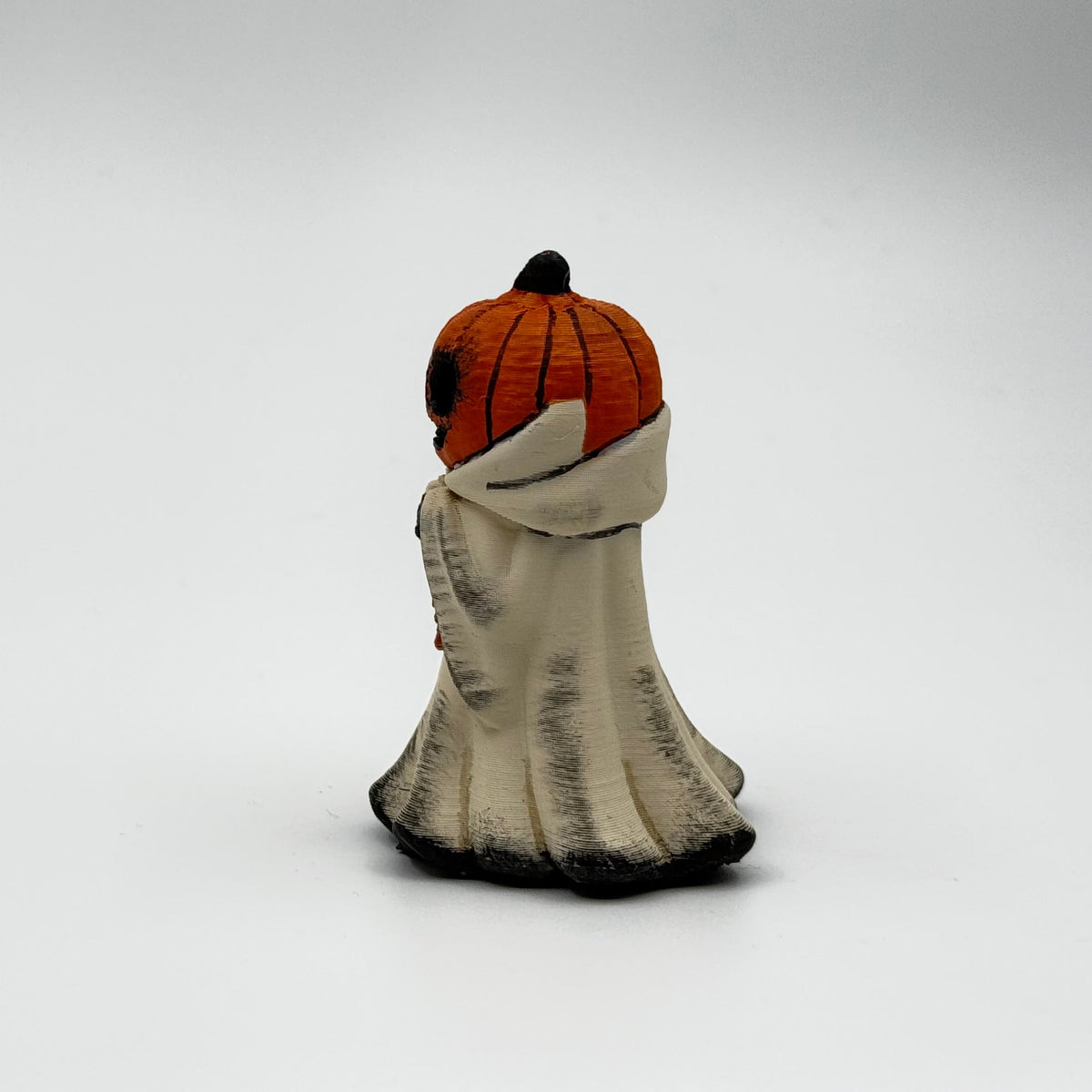 A hand painted folklore figurine made by The Nordic Ghost Merchants in Denmark. The figurine has a carved pumpkin head and is robed in cream cloth while he clutches an autumn leaf. Side view on a grey studio background.