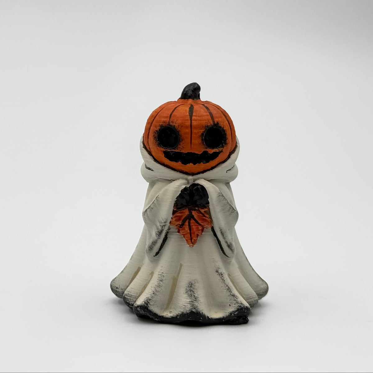 A hand painted folklore figurine made by The Nordic Ghost Merchants in Denmark. The figurine has a carved pumpkin head and is robed in cream cloth while he clutches an autumn leaf. Front view on a grey studio background.