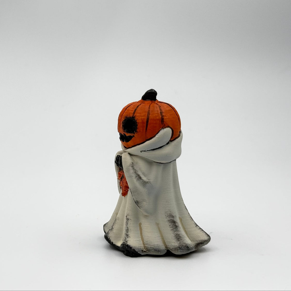 A hand painted folklore figurine made by The Nordic Ghost Merchants in Denmark. The figurine has a carved pumpkin head and is robed in cream cloth while he clutches an autumn leaf. Side view on a grey studio background.