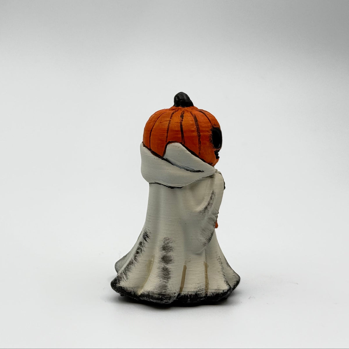 A hand painted folklore figurine made by The Nordic Ghost Merchants in Denmark. The figurine has a carved pumpkin head and is robed in cream cloth while he clutches an autumn leaf. Side view on a grey studio background.