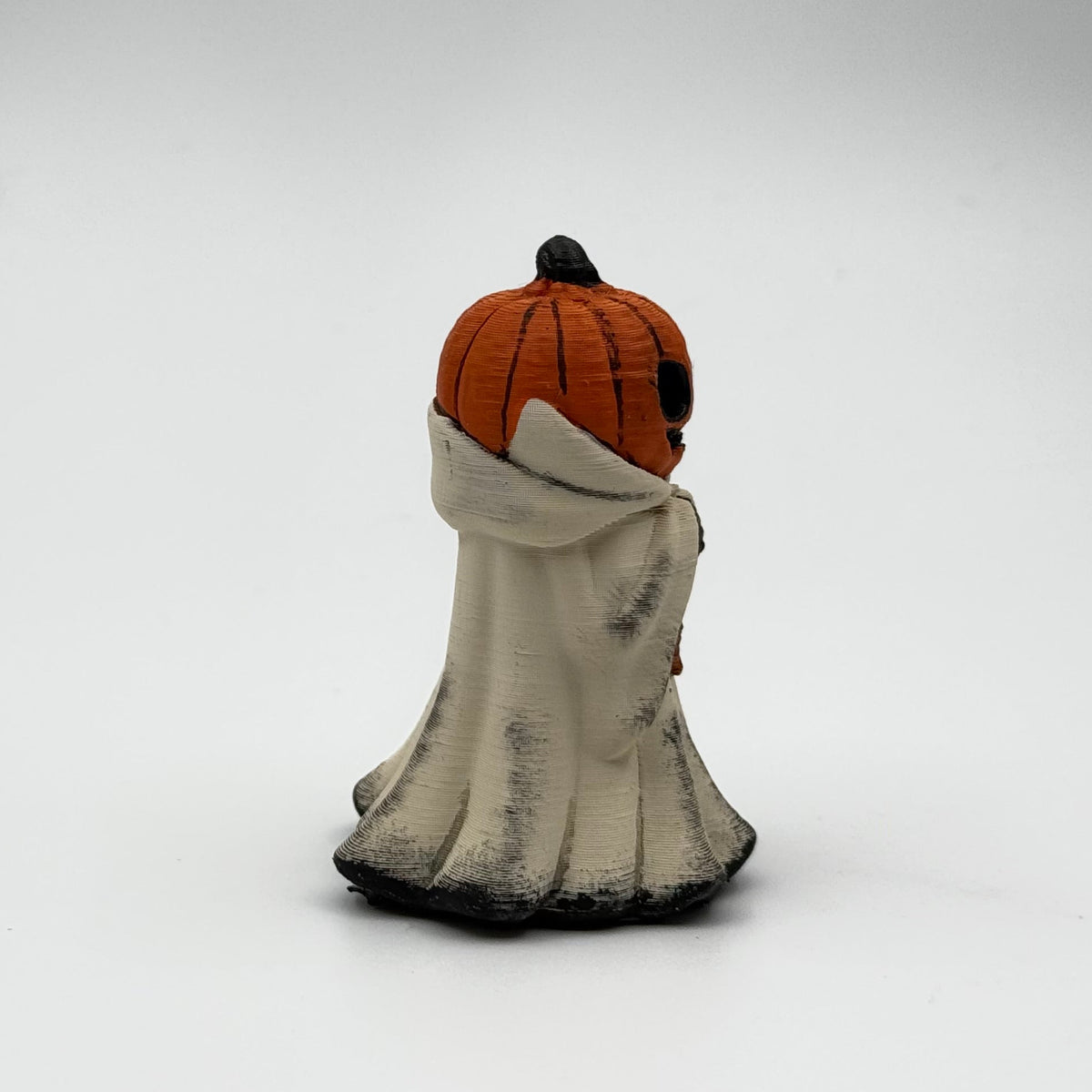 A hand painted folklore figurine made by The Nordic Ghost Merchants in Denmark. The figurine has a carved pumpkin head and is robed in cream cloth while he clutches an autumn leaf. Side view on a grey studio background.