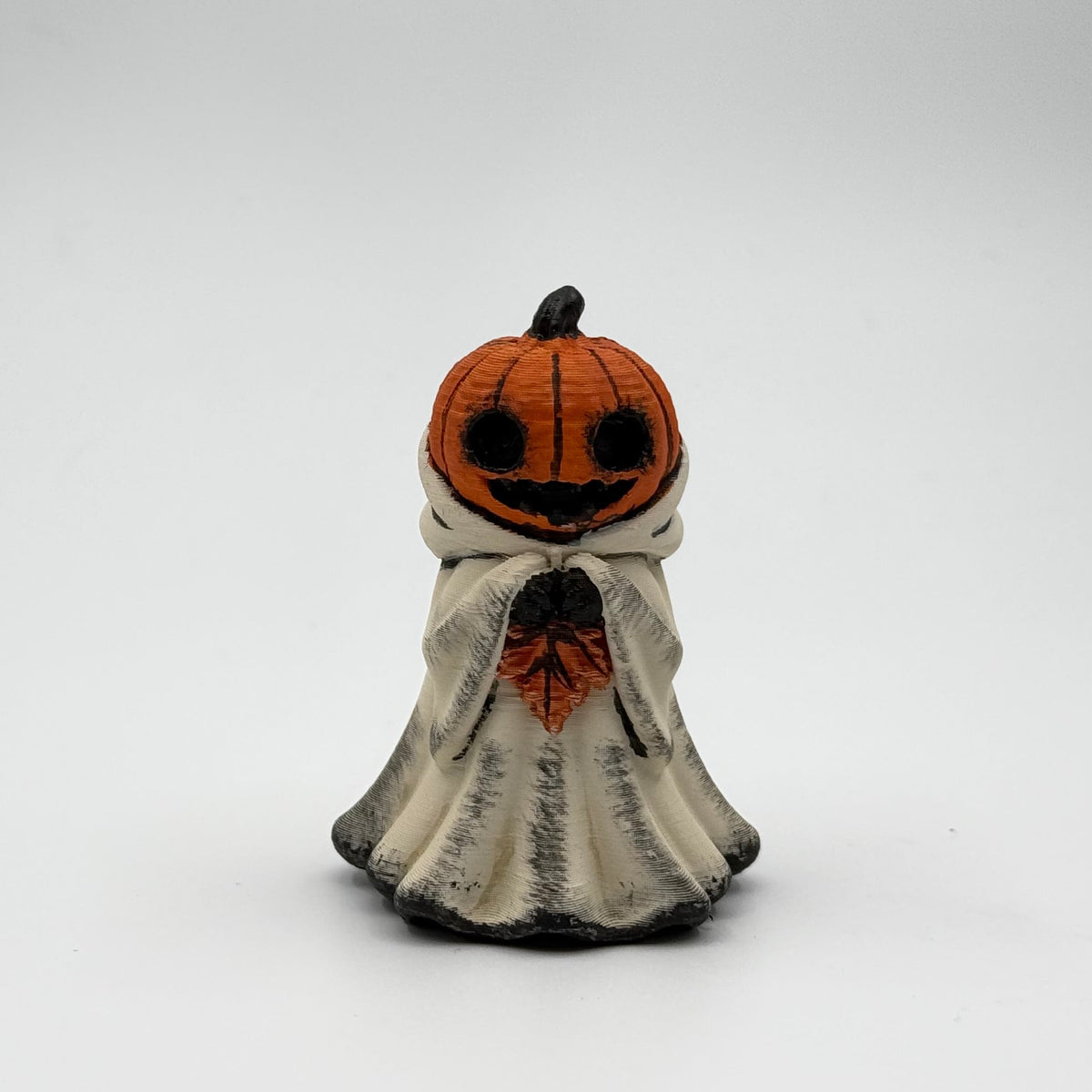 A hand painted folklore figurine made by The Nordic Ghost Merchants in Denmark. The figurine has a carved pumpkin head and is robed in cream cloth while he clutches an autumn leaf. Front view on a grey studio background.