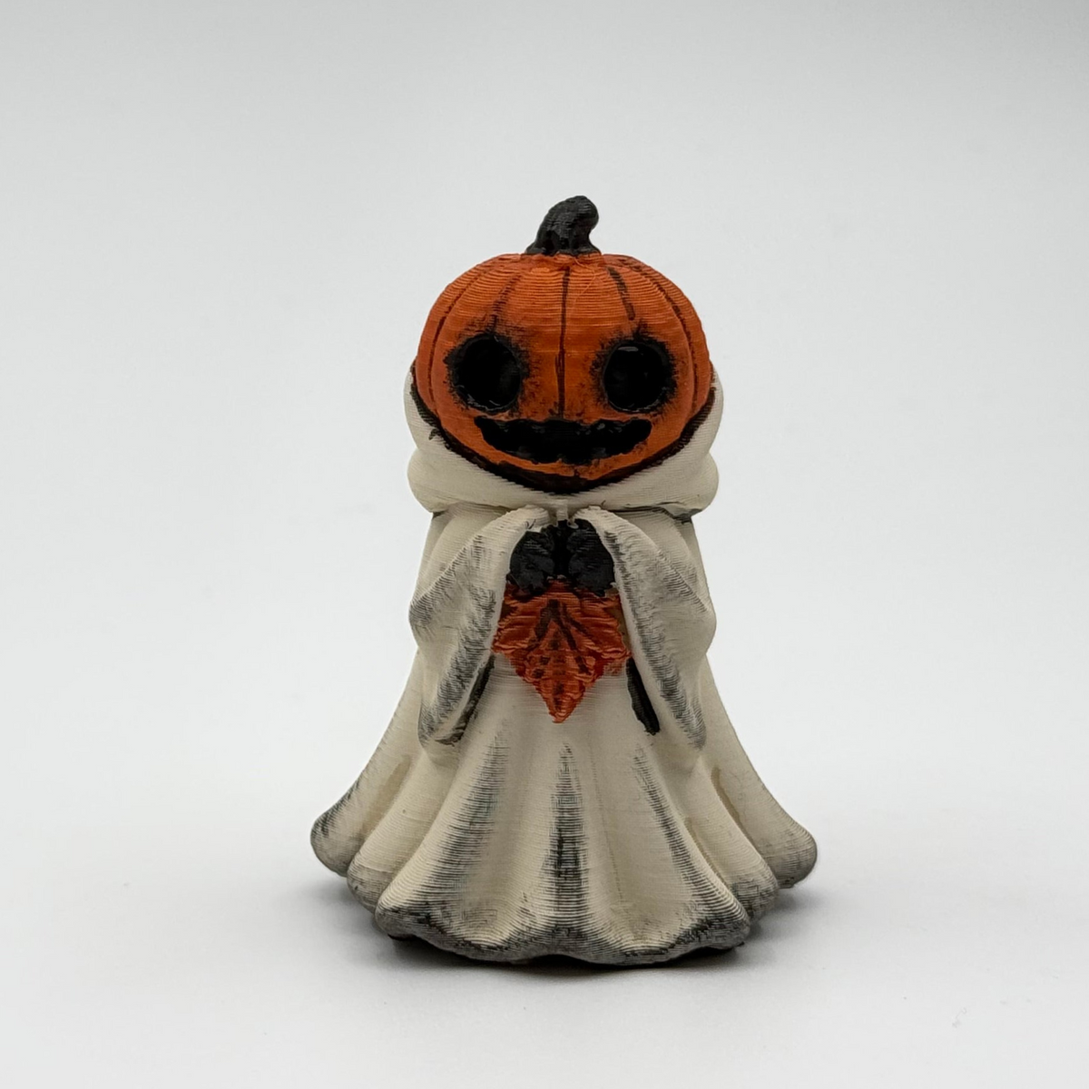 A hand painted folklore figurine made by The Nordic Ghost Merchants in Denmark. The figurine has a carved pumpkin head and is robed in cream cloth while he clutches an autumn leaf. Front view on a grey studio background.