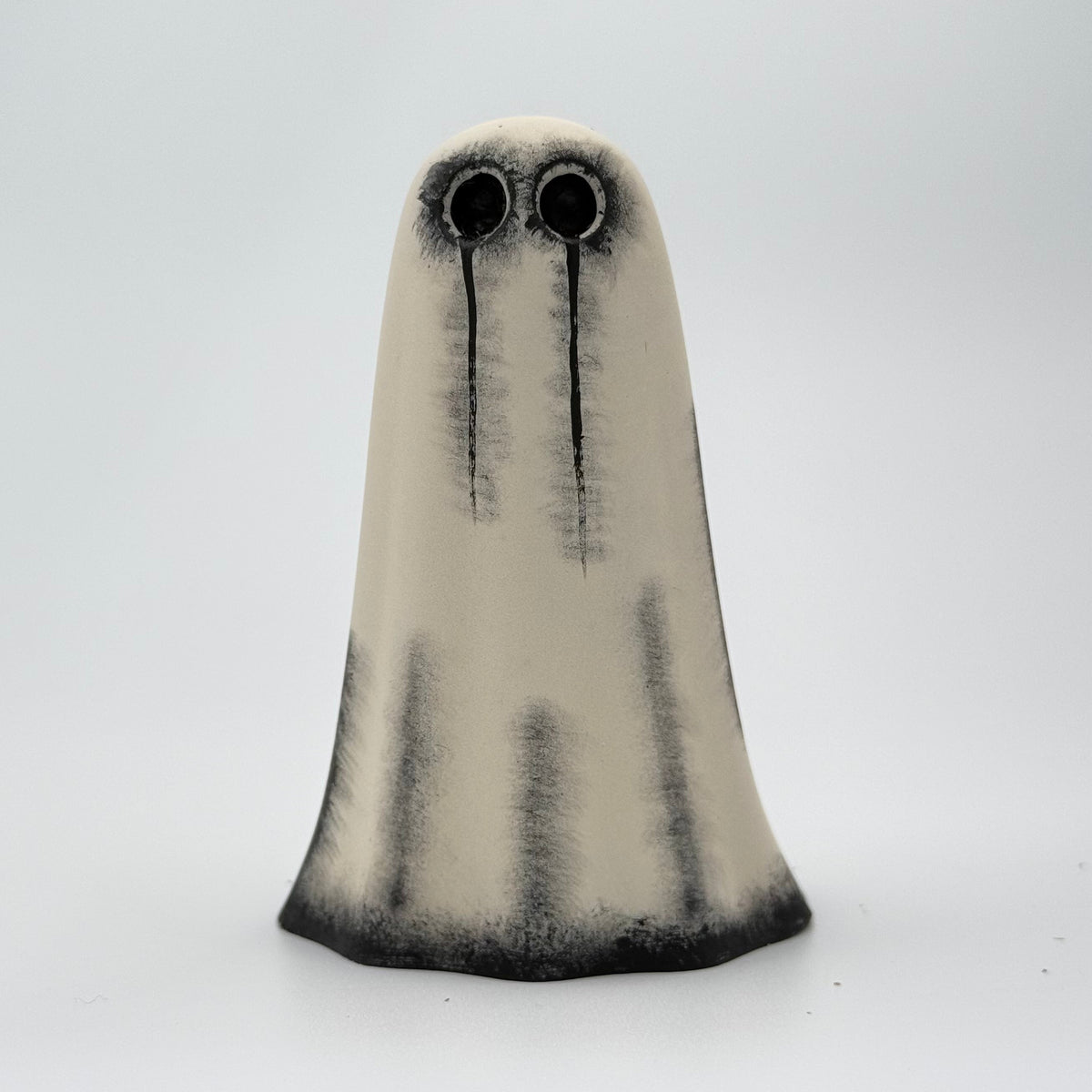 An ivory handmade ghost figurine made by The Nordic Ghost Merchants. The ghost has hand painted black eyes and tear details. Front view on a grey studio background.