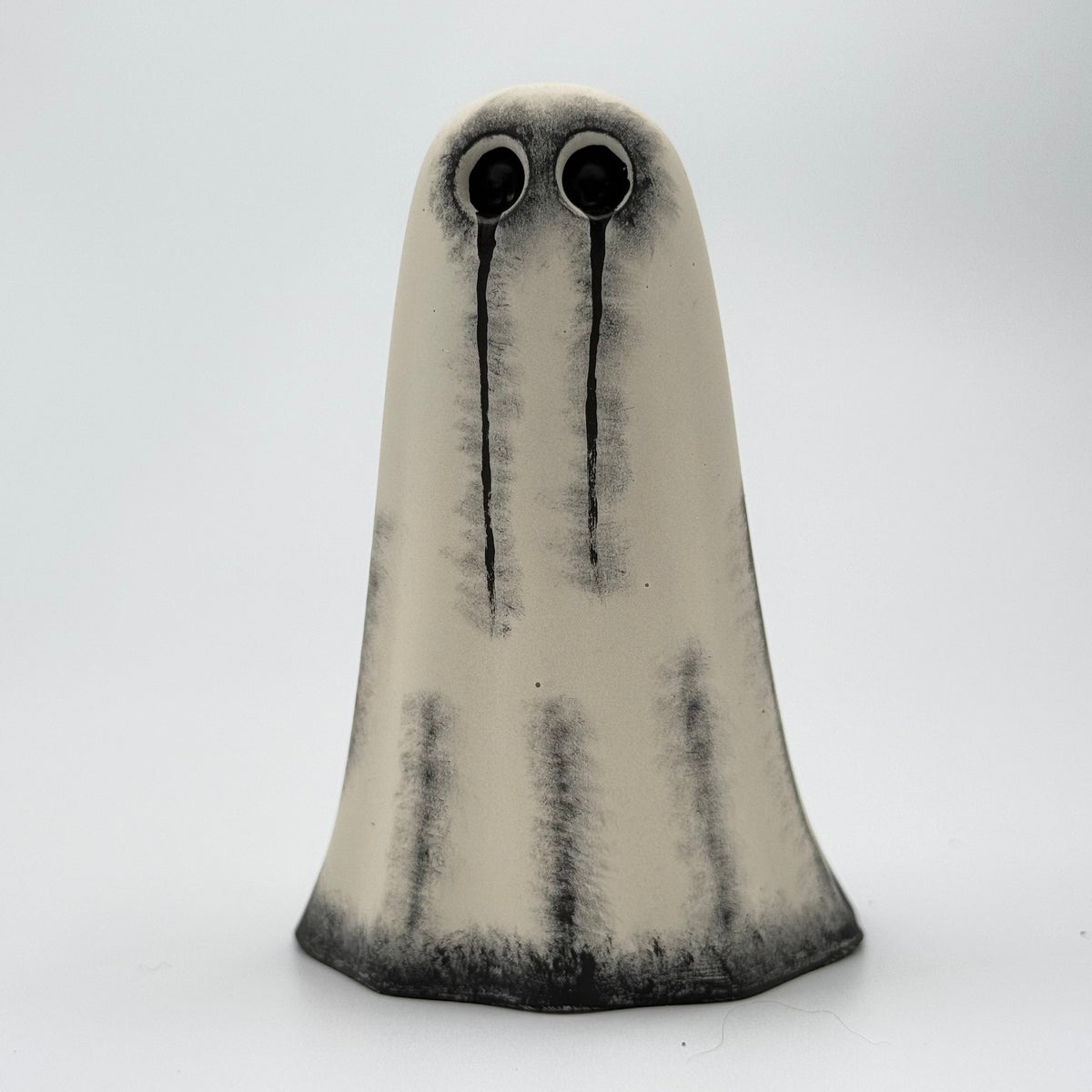A handmade ghost figurine made by The Nordic Ghost Merchants. The body of the ghost is an ivory cream colour with distressed black details around the base and along the folds of the sheet ghost. The ghost has hand painted black eyes with tear stains running downwards. Side view on a grey studio background.