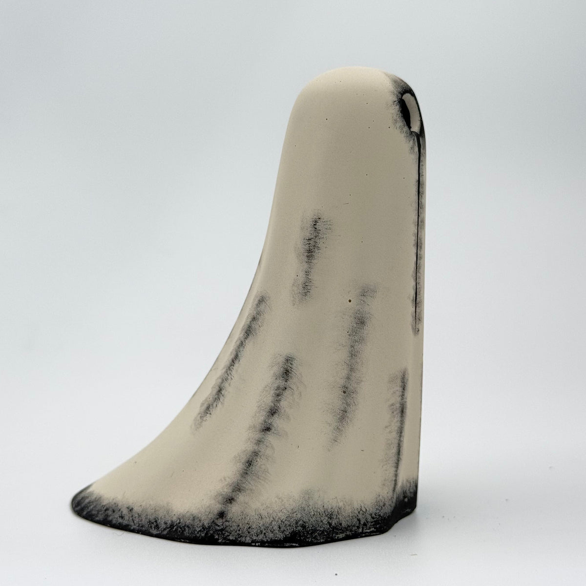 A handmade ghost figurine made by The Nordic Ghost Merchants. The body of the ghost is an ivory cream colour with distressed black details around the base and along the folds of the sheet ghost. Side view on a grey studio background.