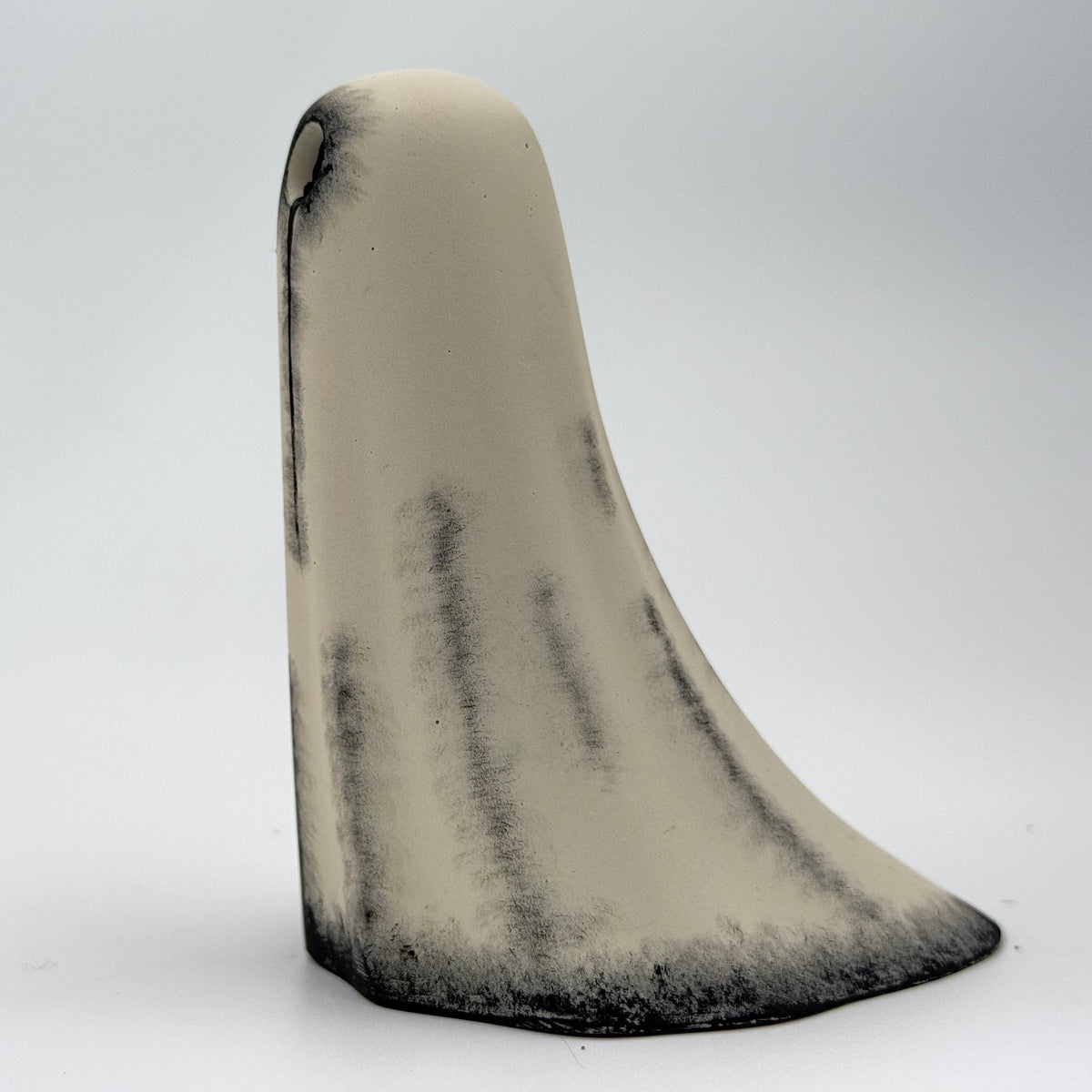 A handmade ghost figurine made by The Nordic Ghost Merchants. The body of the ghost is an ivory cream colour with distressed black details around the base and along the folds of the sheet ghost. Side view on a grey studio background.