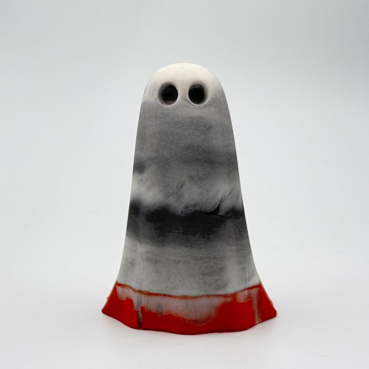 A handmade ghost figurine made by The Nordic Ghost Merchants in Denmark. The body of the ghost is a grey, white and charcoal smoky colour. The bottom centimetre is a dark pumpkin colour. Front view on a grey studio background.