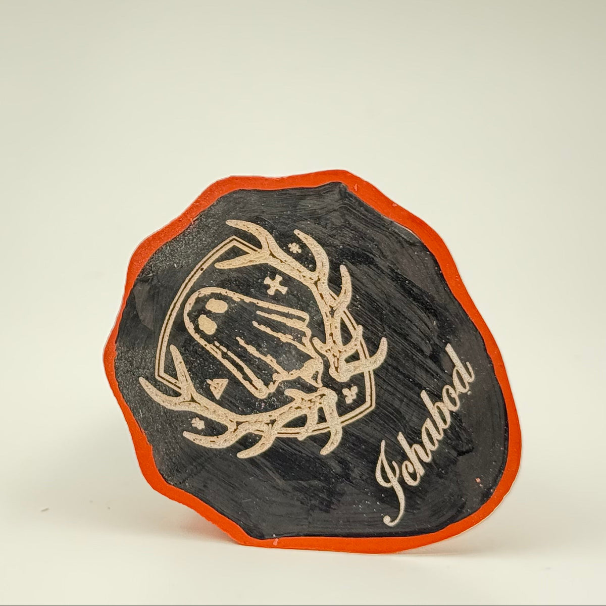 A handmade ghost figurine by The Nordic Ghost Merchants. The view is of the base of the ghost ornament, painted black with an orange border and engraved with the brand logo - a coat of arms with a sheet ghost in the centre.