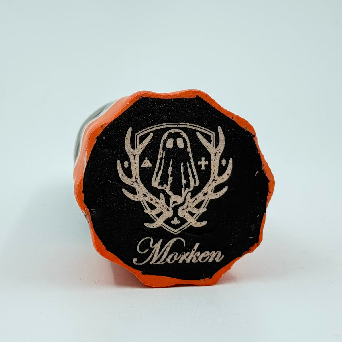 A handmade ghost figurine made by The Nordic Ghost Merchants in Denmark. The view is of the base of the ghost ornament, painted black with an orange border. The base is engraved with the brand logo - a coat of arms with a sheet ghost in the centre. On a grey studio background.