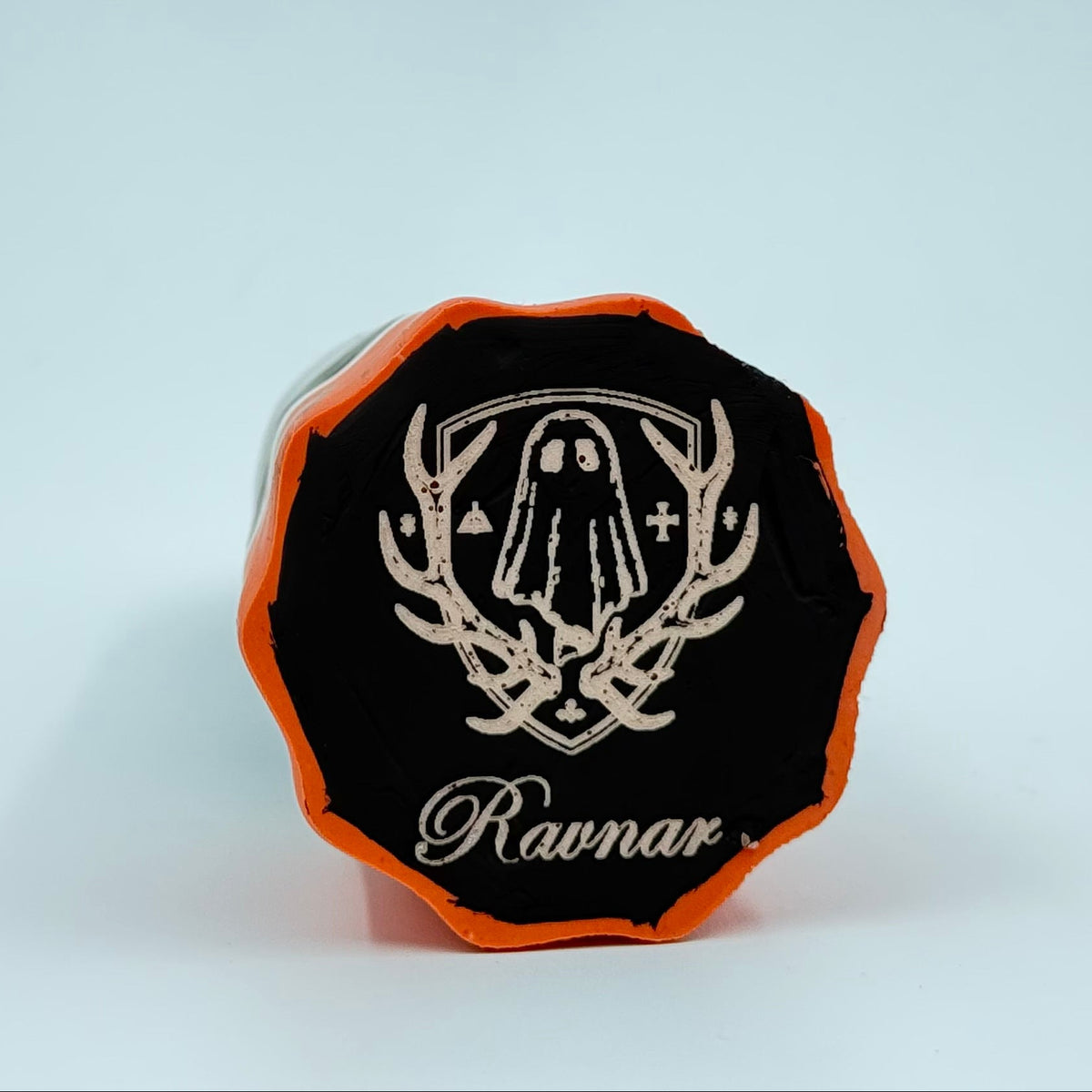 A handmade ghost figurine made by The Nordic Ghost Merchants in Denmark. The view is of the base of the ghost ornament, painted black with an orange border. The base is engraved with the brand logo - a coat of arms, with a sheet ghost in the centre. On a grey studio background.