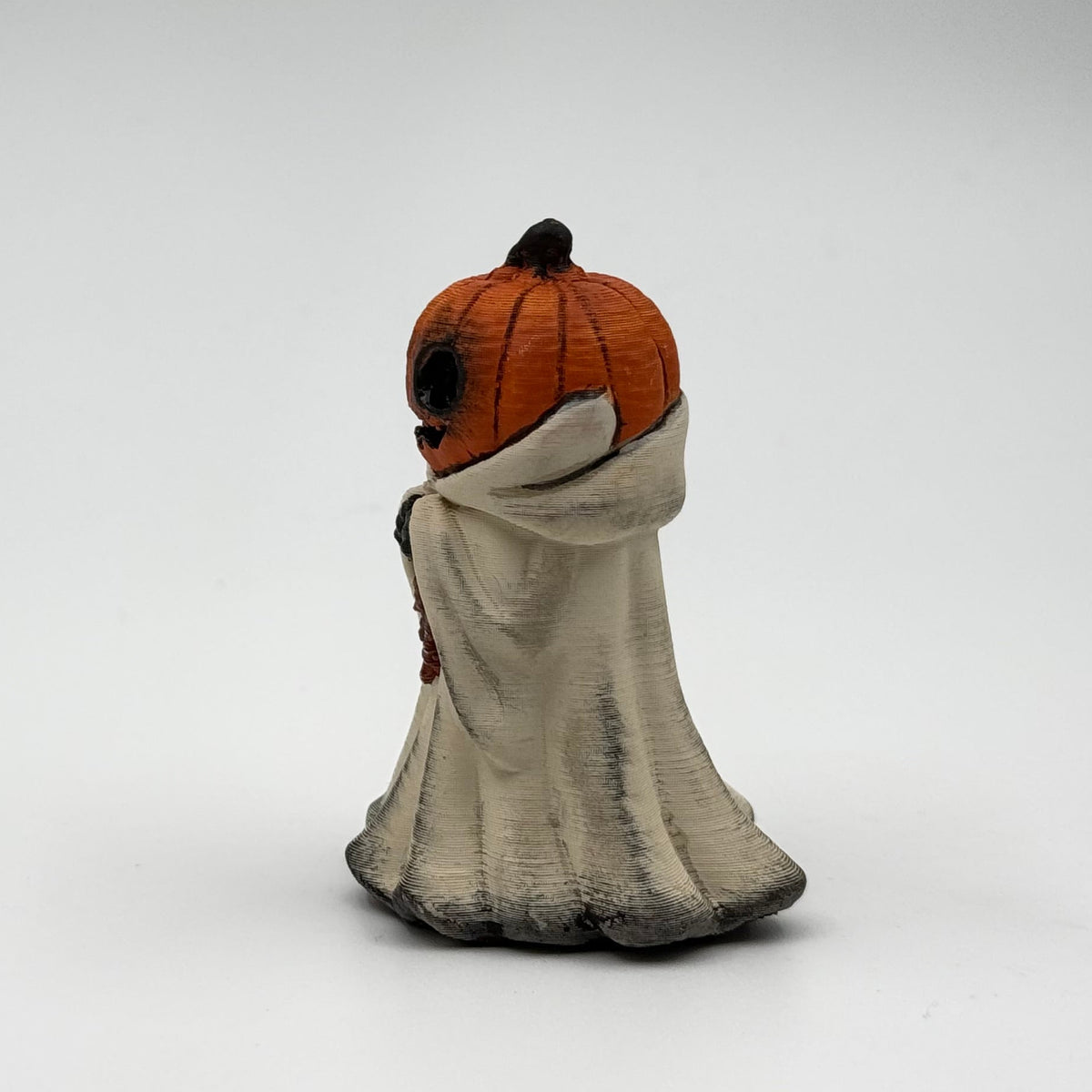 A hand painted folklore figurine made by The Nordic Ghost Merchants in Denmark. The figurine has a carved pumpkin head and is robed in cream cloth while he clutches an autumn leaf. Side view on a grey studio background.
