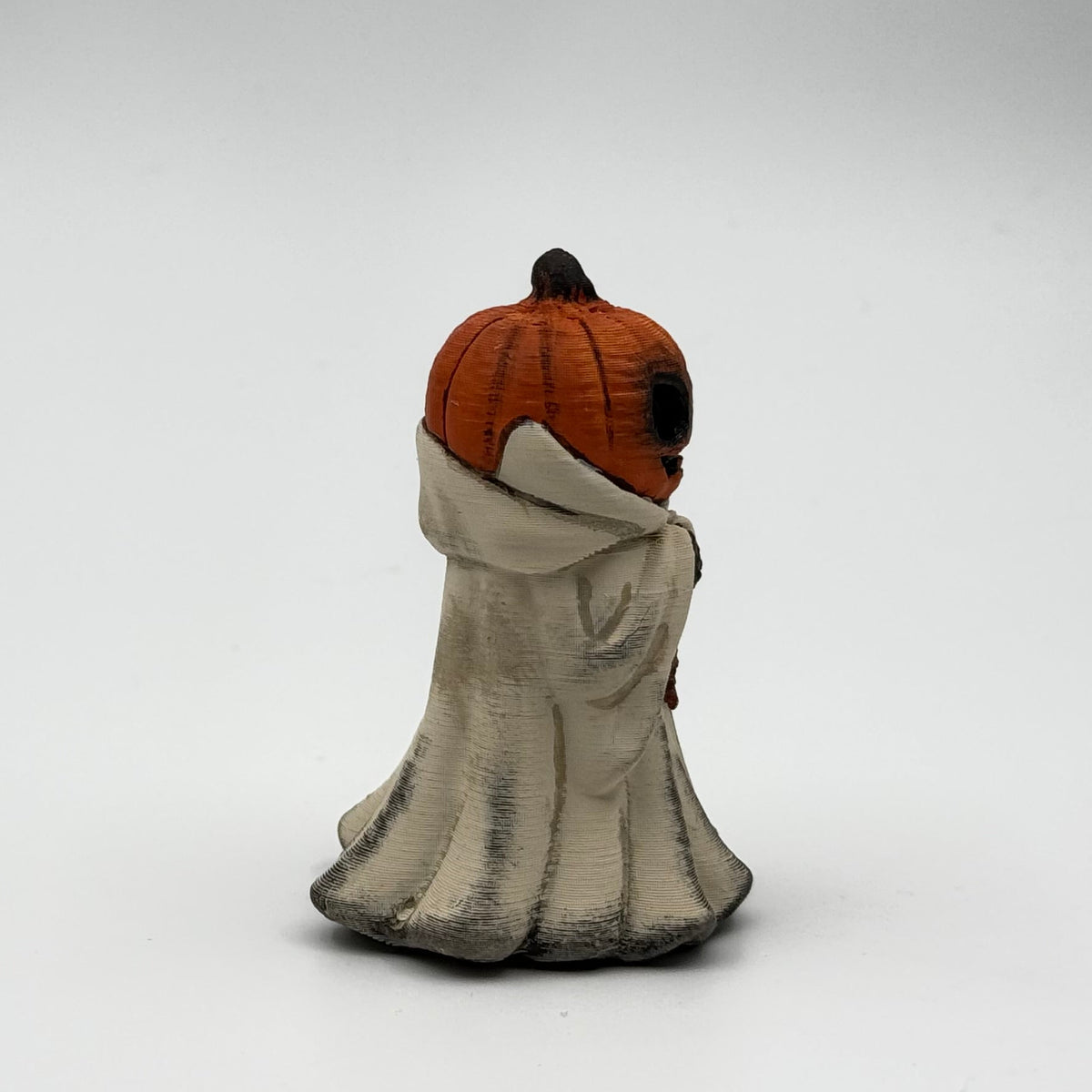 A hand painted folklore figurine made by The Nordic Ghost Merchants in Denmark. The figurine has a carved pumpkin head and is robed in cream cloth while he clutches an autumn leaf. Side view on a grey studio background.